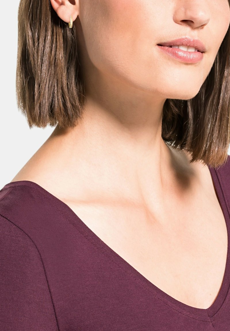 Top a V scollo in tessuto bordeaux scuro, con maniche corte e una superficie liscia. Orecchino a cerchio dorato visibile all'orecchio del modello.