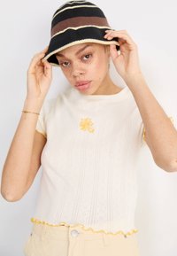 Witte, T-shirt met korte mouwen met een textuurpatroon en gele bloemenborduursels; gerimpelde zoom en manchetten. Zwarte emmerhoed met beige en bruine strepen.