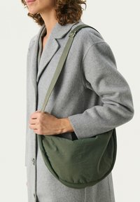 Borsa a tracolla verde in tessuto con una forma morbida e arrotondata. Cinghia regolabile e texture liscia. Indossata sotto un cappotto grigio di lana.