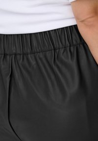 Crne hlače od umjetne kože s elastičnom elastikom u struku, koje imaju glatku teksturu i naboranu tkaninu za udobnost.