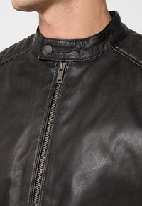 Giacca da moto in pelle nera con colletto rialzato, zip argentata e dettagli trapuntati sulle spalle. Texture liscia con accenti minimi.