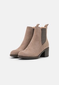 Paire de bottines en daim taupe avec panneaux latéraux élastiques noirs et talons empilés sur fond blanc.