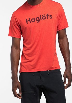 Man wearing a bright red short-sleeve T-shirt with "Haglöfs" logo and black pants with zippered pockets.