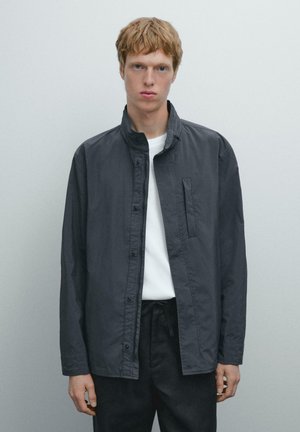 Dark gray lightweight jacket with a stand collar, front button closure, and a single chest pocket. Worn over a white t-shirt.