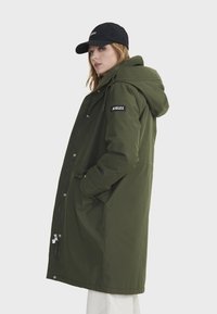 Parka olive vert avec capuche, dotée d'une fermeture éclair frontale et de boutons-pression, ainsi qu'un patch logo noir sur la manche. Texture du tissu lisse.
