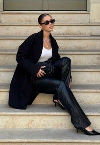 Svart överrock, vit linne och svarta byxor i konstläder. Tillbehör inkluderar liten svart väska och solglasögon. Klackar fullbordar looken.