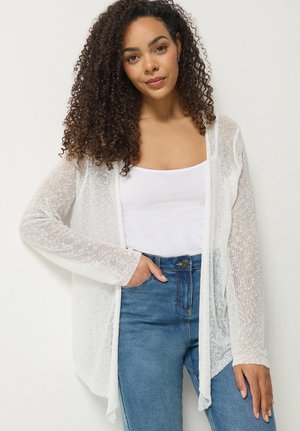 Kvinde med krøllet hår iført en hvid tanktop, gennemsigtig hvid cardigan og blå jeans, stående med den ene hånd i lommen.