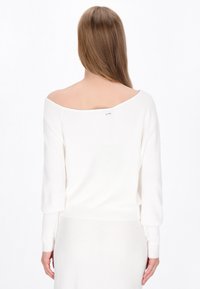 Suéter blanco de punto con un diseño off-the-shoulder, mangas largas y puños acanalados. Presenta una pequeña etiqueta metálica en la parte posterior.