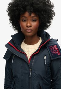 Granatowa ocieplana kurtka z wysokim kołnierzem, czerwonymi akcentami na suwakach oraz naszywką z napisem "SUPERDRY MNTN". Posiada kieszenie na zamek.