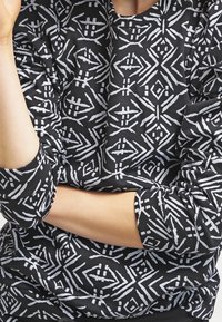 Jersey negro de manga larga con un patrón geométrico blanco. Puños y dobladillo acanalados, tejido suave con un acabado texturizado.