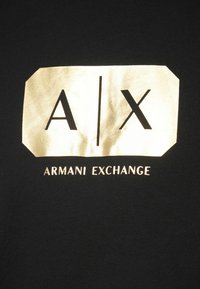 Mustad must T-särk, millel on suur kuldne ristkülikukujuline logo "A/X" ja allpool valge tekst "ARMANI EXCHANGE".