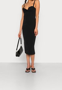 Svart figurnära klänning med hjärtformad urringning och spagettiband, tillsammans med en svart texturerad handväska och platta sandaler.