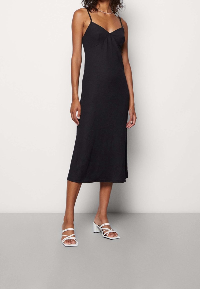 Abito midi nero senza maniche con dettaglio a nodo sul davanti, texture liscia e spacchi laterali; abbinato a sandali bianchi con tacco largo.