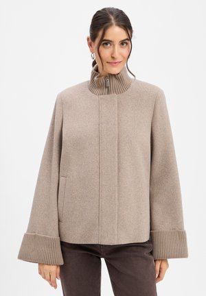 Frau trägt eine beige Wolljacke mit hohem geripptem Kragen und breiten gerippten Bündchen, steht vor einem einfarbigen weißen Hintergrund.