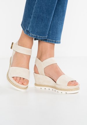 Fødder iført beige kilehælssandaler med ankelremme kombineret med blå cropped denimjeans på hvid baggrund.