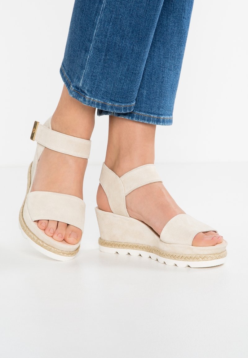 Pieds portant des sandales compensées beiges avec des brides à la cheville associées à un jean denim bleu raccourci sur fond blanc.