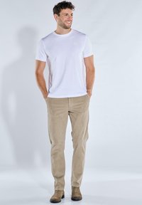 Weißes T-Shirt mit Rundhalsausschnitt, schmal geschnittene beigefarbene Cordhosen und braune Stiefeletten. Das Model steht mit den Händen in den Taschen vor einem hellen Hintergrund.