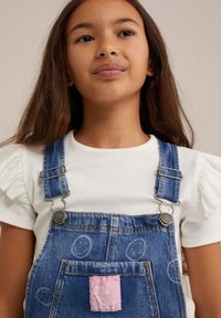 Pantalones de mezclilla azul con herrajes plateados, tirantes ajustables, bolsillo frontal con un parche rosa con texto, acompañados de una camiseta blanca de canalé.