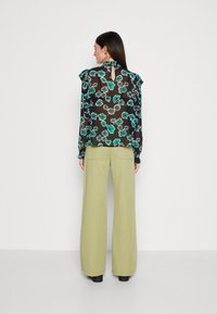 Schwarzbluse mit langen Ärmeln, tealfarbenem und pinkem Blumenmuster, mit Rüschensäumen und Schlitz im Rücken, kombiniert mit weit geschnittenen, olivgrünen Hosen.