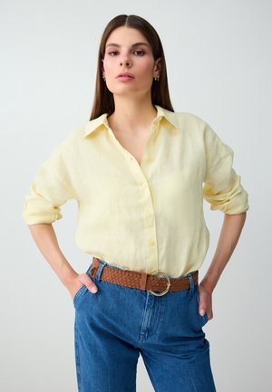 Vrouw met lang bruin haar die een lichtgele blouse met knopen draagt, blauwe spijkerbroek en een bruine gevlochten riem, poseert met handen in de zakken.