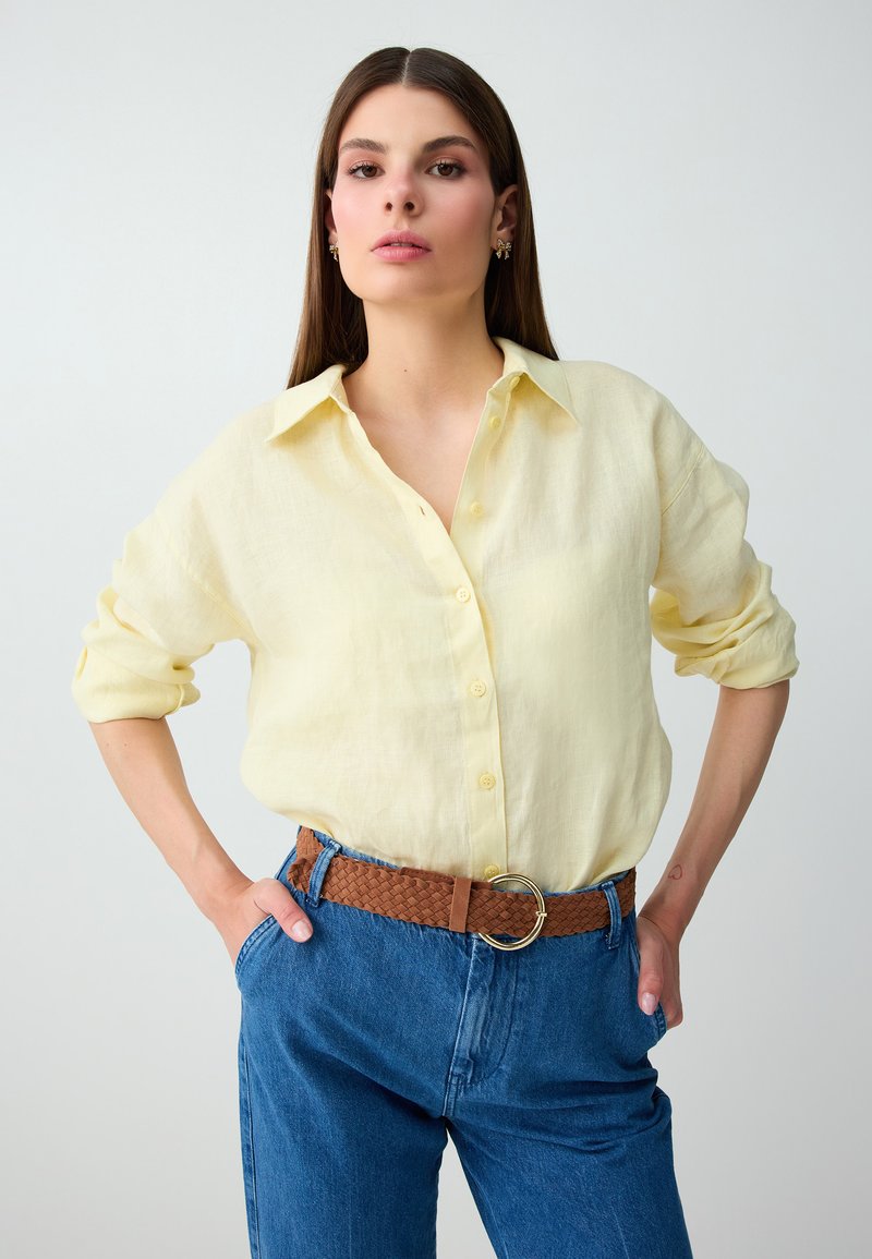 Femme aux longs cheveux bruns portant une chemise jaune clair boutonnée, un jean bleu et une ceinture tressée marron, pose les mains dans les poches.