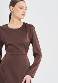 Braunes langärmliges Kleid mit quadratischem Ausschnitt, das eine glatte, satinähnliche Textur und eine maßgeschneiderte Silhouette aufweist, die durch strukturierte Schultern betont wird.