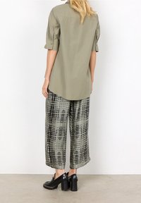 Camicia verde oliva a maniche corte con vestibilità ampia e orlo posteriore. Abbinata a pantaloni larghi grigi con motivi e linee astratte nere. Scarpe con piattaforma nere.