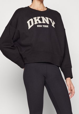 Person iført en sort oversized sweatshirt med teksten "DKNY New York" og sorte tætsiddende leggings mod en enkel lys baggrund.