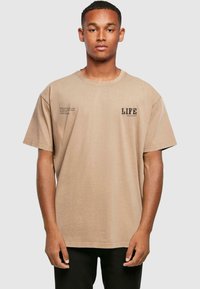 Beige oversized t-shirt i bomuld med korte ærmer, med trykt tekst på venstre bryst og siden. Rund halsudskæring, afslappet pasform.