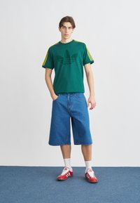 Jauns vīrietis zaļā Adidas T-kreklā, plašās zilās džinsa šortos, baltās zeķēs un sarkanos Adidas sporta apavos stāv uz zilas paklājiņa.