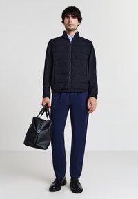 Homme portant une veste matelassée bleue marine et un pantalon, tenant un sac à main en cuir noir, se tenant sur un fond clair.