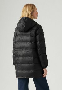 Chaqueta de plumas negra con acolchado horizontal, capucha y mangas largas. Acabado brillante y puños elásticos. Vista desde atrás.