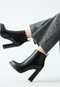 Stivaletti alla caviglia in pelle nera con tacco alto a blocco, con cerniera laterale e dettaglio ad anello, abbinati a jeans grigi corti.