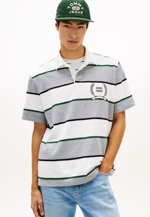 Giovane uomo che indossa un berretto verde Tommy Jeans, una polo a righe grigie e bianche e jeans azzurri, in piedi con una mano in tasca.