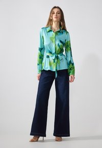 Cămașă albastră deschis cu model în acuarelă verde, cu brâu la talie, asortată cu pantaloni jeans largi de culoare închisă și tocuri bej.