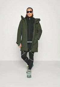 Olivgrön parkajacka med huva, med en frontdragkedja, två sidofickor, lagd över en svart zip-up-tröja och svarta leggings.