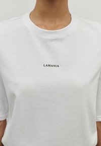 Balta medvilnės marškinėliai trumpomis rankovėmis ir apvaliu kaklu. Priekyje, centre, yra juodais lankiniais užrašytas tekstas "LAMANIA". Lygus paviršius.
