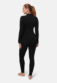 Frau steht barfuß mit zurückgebundenen Haaren, trägt ein eng anliegendes schwarzes Langarmoberteil und schwarze Leggings, steht mit dem Rücken zu einem schlichten Hintergrund.