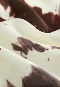 Gros plan d'un tissu lisse avec un fond blanc et des taches brunes irrégulières, montrant des plis doux et une texture.