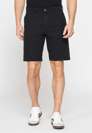Uomo con pantaloncini neri fino al ginocchio e scarpe da ginnastica bianche, in piedi con le gambe leggermente divaricate su uno sfondo chiaro e uniforme.