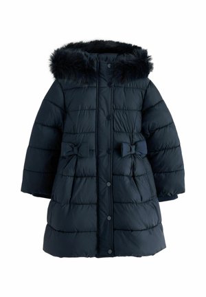 Ung pige iført en sort dunjakke med pels hætte, hvid skjorte, vidde jeans og hvide sneakers, der står mod en beige væg.