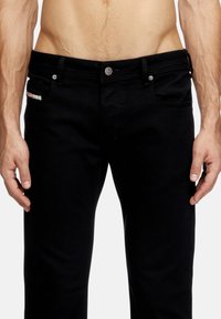 Männlicher Torso, der schwarze Jeans mit Knopfverschluss trägt, sichtbare Gürtelschlaufen, und die Hände entspannt an den Seiten vor einem weißen Hintergrund.