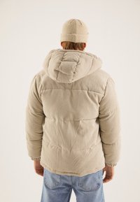 Doudoune en velours côtelé beige avec capuche, présentant une texture côtelée et des poignets élastiques, portée avec un bonnet assorti en tricot. Pantalon en jean bleu visible.