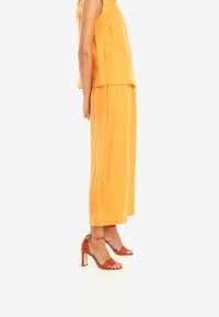 Orange linne ärmlös topp i kombination med hög midja, vida byxor; prydd med orange klacksandaler med remmar. Slät textur överlag.