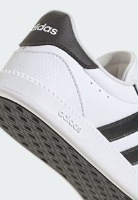Zapatilla blanca con acentos negros, con una superficie texturizada, perforaciones y suela de goma con un patrón de cuadrícula. Logotipo negro en la parte trasera.