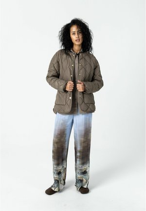 Frau mit lockigem Haar, die eine gesteppte braune Jacke, ein braunes Hemd und weite Hosen mit einem blau-braunen abstrakten Muster trägt, steht vor einem weißen Hintergrund.