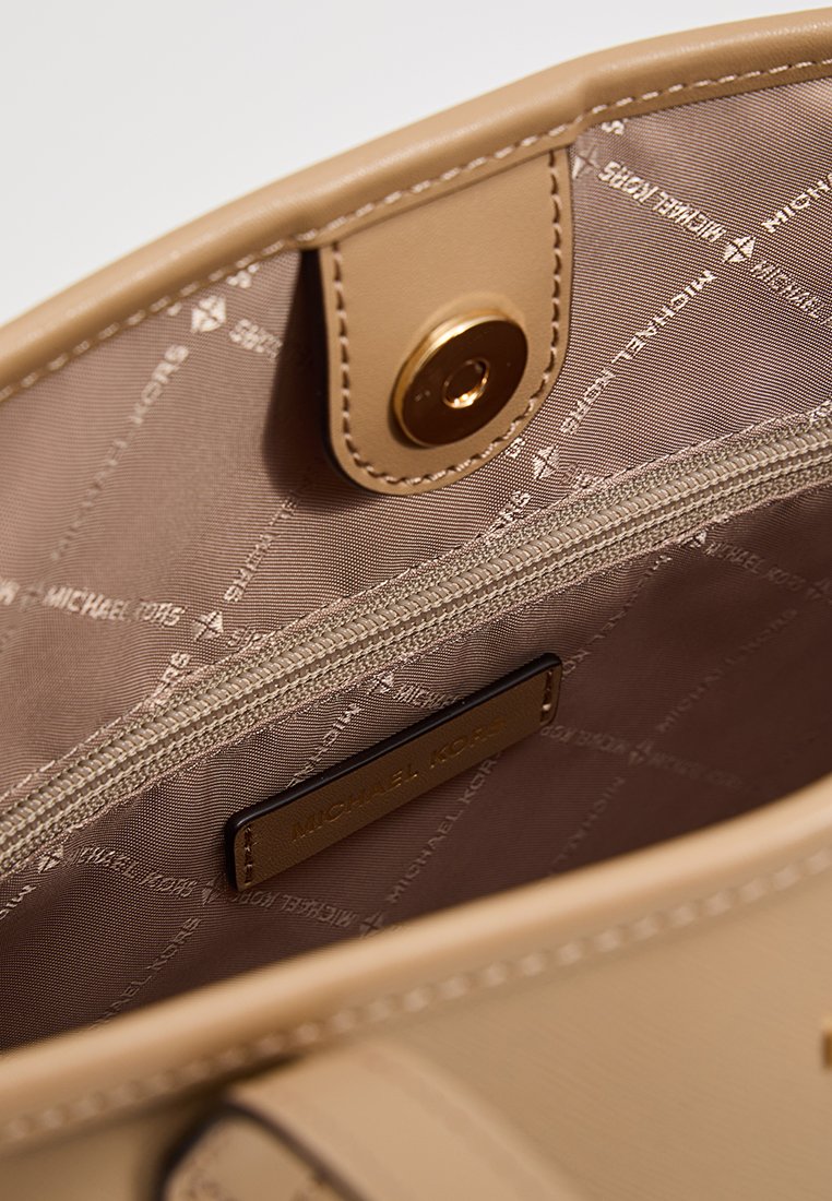 Interno di una borsa a tracolla in pelle beige, con una fodera color tan che presenta il logo "Michael Kors" ripetuto, una tasca con zip e un dettaglio di chiusura a scatto dorato.