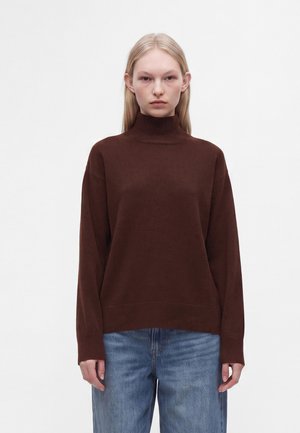 Junge Person mit glatten blonden Haaren, die einen dunkelbraunen Rollkragenpullover und blaue Jeans trägt, steht vor einem schlichten Hintergrund.