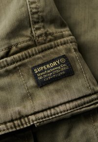 Tela verde oliva con una textura suave, con una etiqueta negra que lleva "SUPERDRY" y detalles de fabricación impresos en amarillo y blanco.