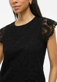 Top in pizzo nero con maniche corte e scallop. Presenta un intricato motivo floreale e una finitura testurizzata. Collo rotondo.